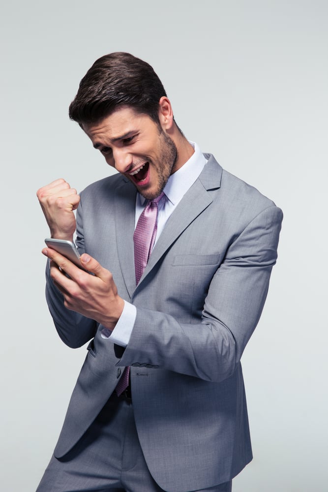 Happy businessman holding smartphone and celebrating his success over gray background